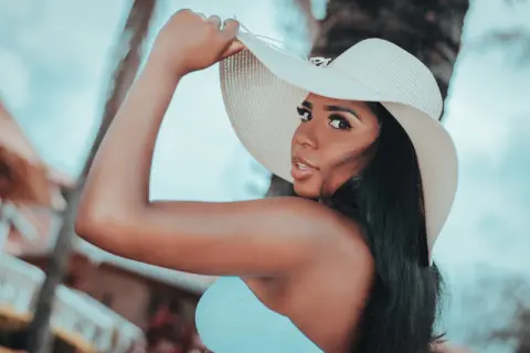 Beautiful woman in a light blue top and wide-brimmed sun hat posing outdoors in a tropical setting.