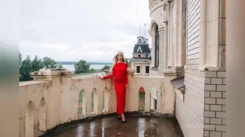 Elegant woman in a red evening gown standing on a stone balcony overlooking a scenic lake view.