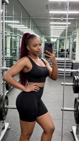 Fit Black woman in black gym outfit taking a mirror selfie in a modern fitness center.