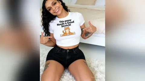 BiancaBell smiles while wearing a white cat-print crop top and black denim shorts, pointing to the text on her shirt while sitting on a white rug.
