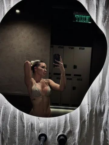 Young woman in a beige bikini taking a mirror selfie in a moody, dimly lit restroom setting.