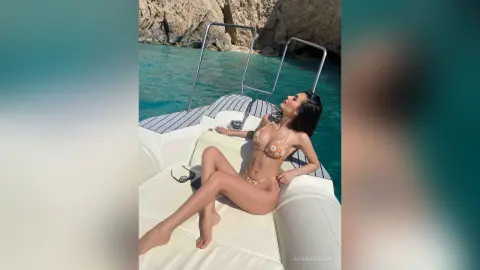 Woman in floral bikini relaxing on a white boat deck in turquoise ocean water near rocky cliffs.
