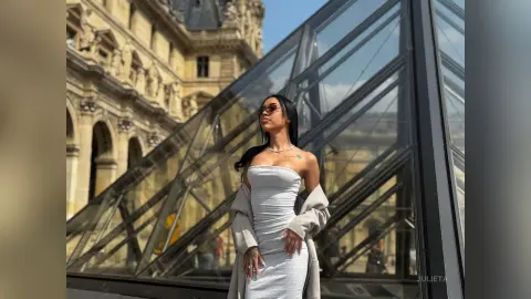 Stylish woman in a grey dress and sunglasses posing by the Louvre Pyramid in Paris under bright sunlight.