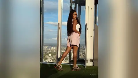 Stylish woman in a white tank top and pink skirt posing by a skyscraper window overlooking a sunny city skyline.