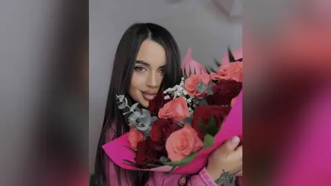 Smiling young woman with long dark hair holding a large bouquet of pink and red roses in a soft romantic setting.