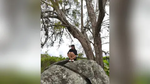 SamaraBert sits on a large rock outdoors, wearing a black t-shirt, white shorts, and a black cap, surrounded by tall trees.