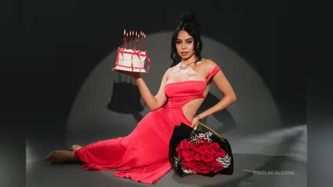 Glamorous woman in a red evening gown holding a lit birthday cake in a moody studio setting.