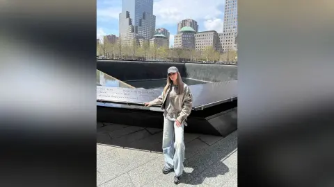 Stylish woman in a beige jacket and light jeans standing by a memorial in a sunny urban city setting.