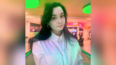 Young woman with long dark hair wearing a white button-down shirt in a brightly lit indoor setting.