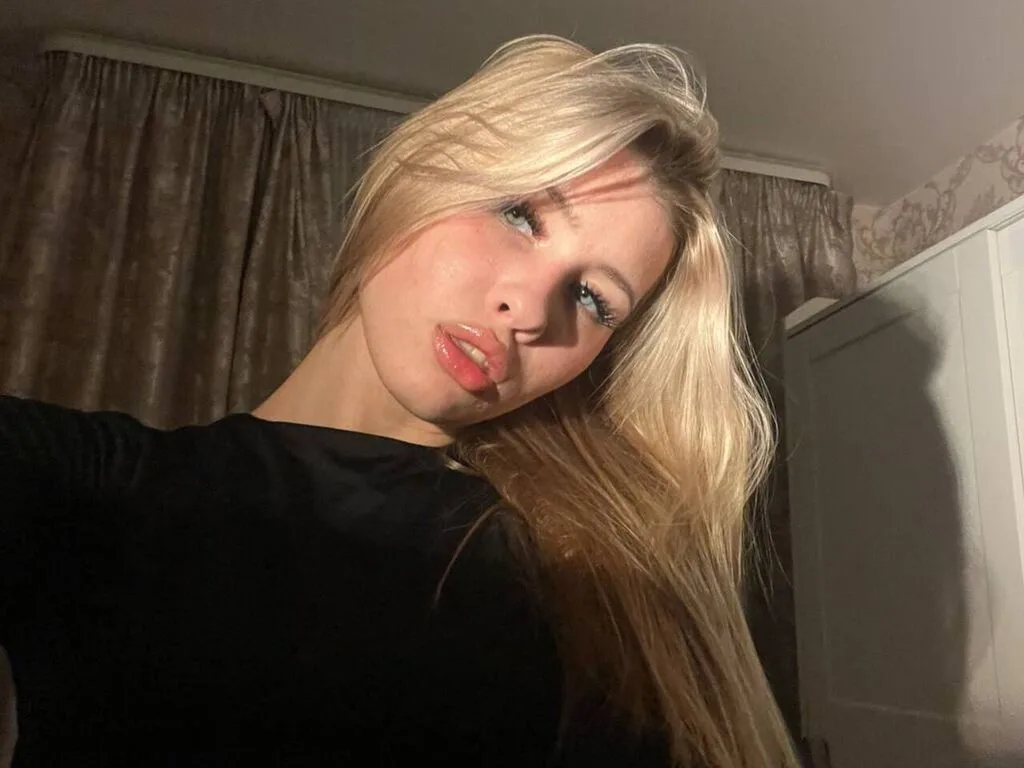 RowanBellamy poses with long blonde hair and light blue eyes, wearing a black top against a backdrop of brown curtains.