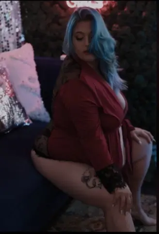 Curvy woman with blue hair in a red silk robe relaxing on a velvet sofa in a moody, glamorous setting.