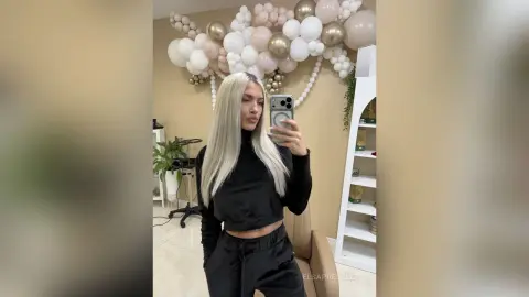 Blonde woman in a black long-sleeve crop top posing in a room decorated with white and gold balloons.