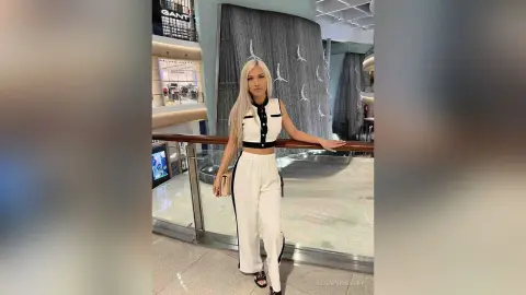 Blonde woman in a chic white two-piece outfit posing confidently by a modern indoor water feature.