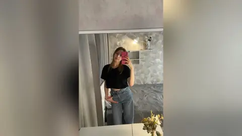 Young woman in a black crop top and blue jeans taking a mirror selfie in a modern, bright bedroom.