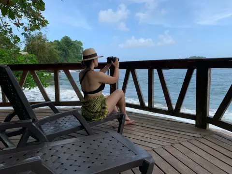 Woman in a sun hat and yellow patterned skirt sitting on a wooden deck overlooking the ocean waves.