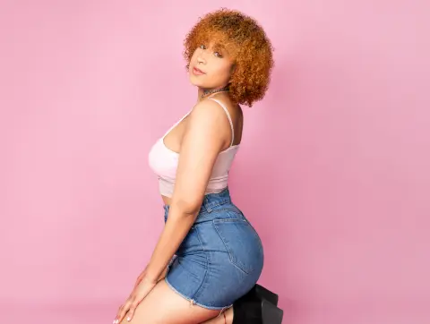 Curvy woman with curly hair in a white tank top and denim shorts posing against a pink background.