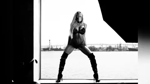 LeslyAnderson stands naked in black lace lingerie and thigh-high boots against a bright waterfront background.