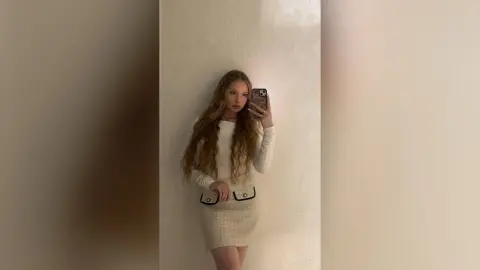 ViolaFausel poses in a white long-sleeve top and matching skirt, holding a phone for a mirror selfie against a plain white wall.