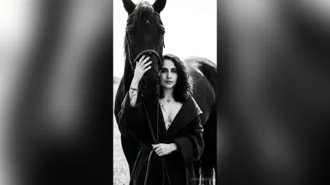 Moody black and white portrait of a woman in a dark coat leaning against a large horse in a field.