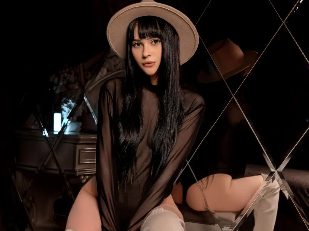 Stylish woman in a black sheer top and wide-brimmed hat posing in a moody, dark mirrored setting.