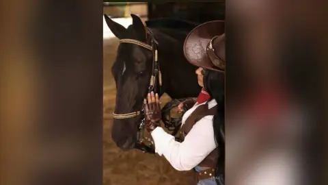 Cowgirl in western attire bonding with a dark brown horse in a rustic stable setting.
