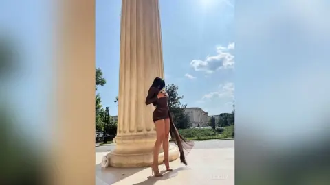 NicoleBella stands outdoors in a short brown dress, posing next to a large stone column under a bright sky.