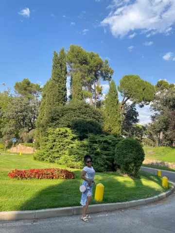 Smiling woman in a white t-shirt and denim skirt standing in a sunny green park with tall trees.