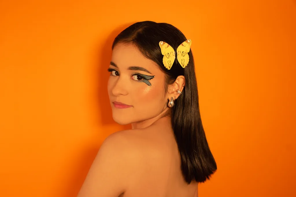 SophieeJacobs poses with a side profile, showing her long dark hair and a yellow butterfly clip against a bright orange background.
