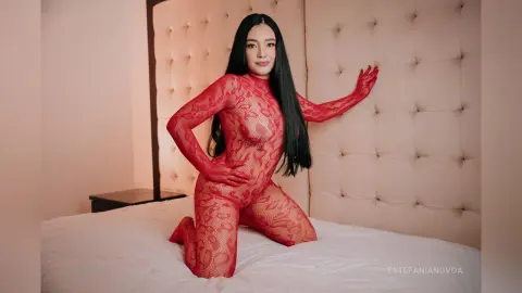 Young woman with long black hair wearing a red lace bodysuit posing on a white bed in a bright bedroom.