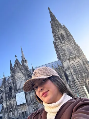 Young woman in a white turtleneck and brown jacket smiling in front of the historic Cologne Cathedral.