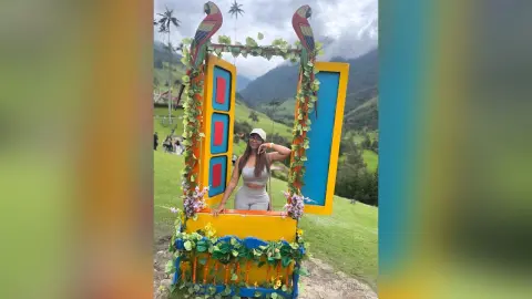 Woman in casual summer outfit posing inside a colorful decorative frame in a lush green mountain landscape.