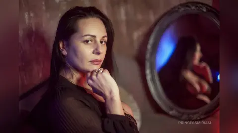 Moody portrait of a woman in a black blouse sitting near a vintage mirror in soft, dim lighting.