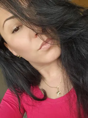 FoxyKara posing for a selfie with her long black hair falling over her face, wearing a pink top and a gold star necklace.