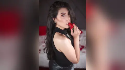 SamanthaHarvey licks a fresh strawberry while wearing a black sleeveless top and looking at the camera.