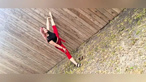 Young woman in black crop top and red leggings stretching outdoors on a wooden deck near a grassy slope.