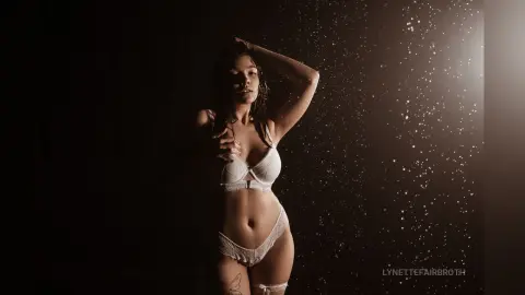 Moody portrait of a woman in white lace lingerie posing under falling water droplets against a dark background.