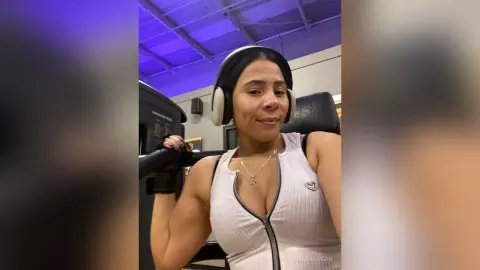 Smiling woman in white ribbed gym tank top wearing headphones during a workout in a modern fitness center.