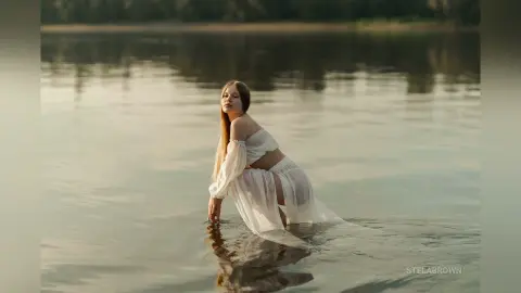 StelaBrown poses in a white sheer dress while kneeling in shallow lake water.