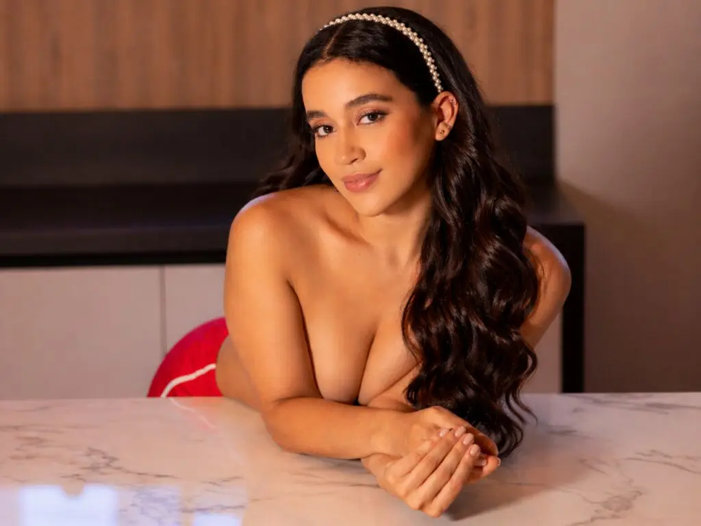 Young woman with long wavy hair and pearl headband leaning on a marble table in a warm indoor setting.