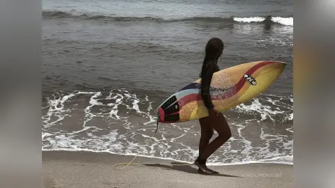 Woman in black wetsuit carrying a colorful surfboard along a wavy beach at sunset.