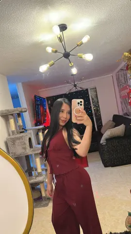 Young woman in a burgundy jumpsuit taking a selfie in a cozy, dimly lit living room.