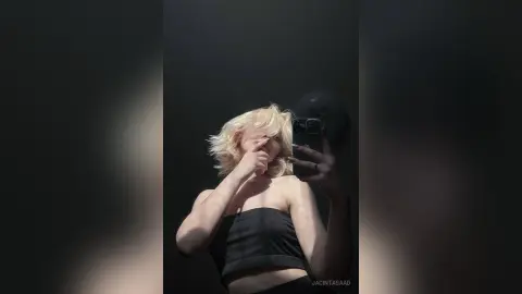 Blonde woman in a black tube top taking a moody selfie against a dark background.