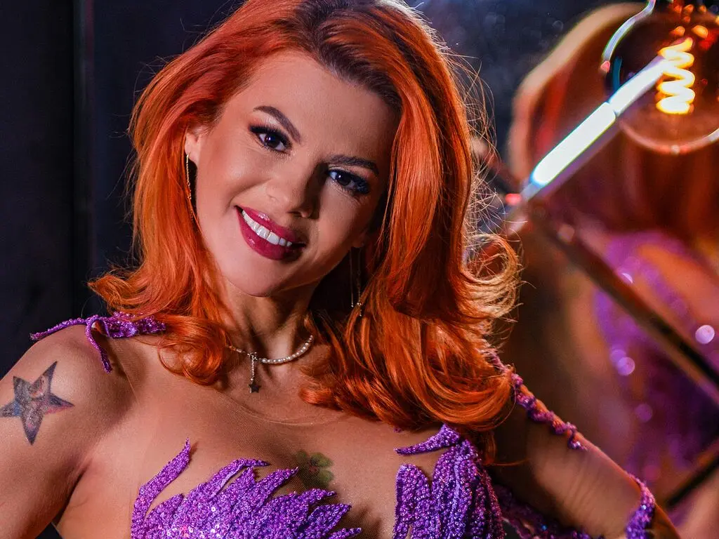 Smiling redhead woman in a sparkly purple sequin outfit posing in a moody, dimly lit setting.