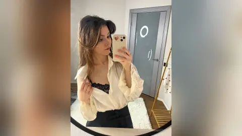 Young woman in a white silk blouse and black skirt taking a mirror selfie in a bright modern room.