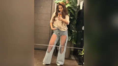 Woman in beige knit top and ripped wide-leg jeans taking a mirror selfie in a modern room.