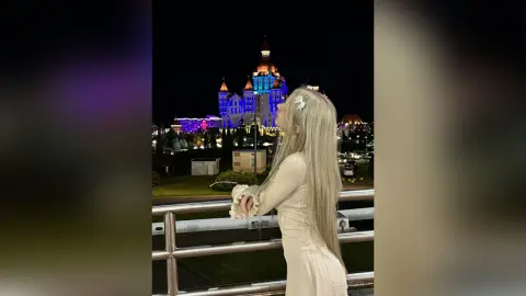 Blonde woman in a white long-sleeve dress gazing at a glowing fairytale castle at night.