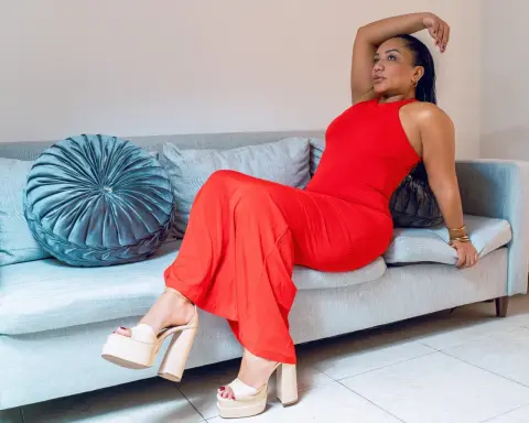 Elegant woman in a vibrant red jumpsuit relaxing on a grey sofa in a bright, modern living room.