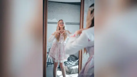 Young woman in a floral white dress making a heart gesture in a mirror, creating a dreamy and romantic mood.