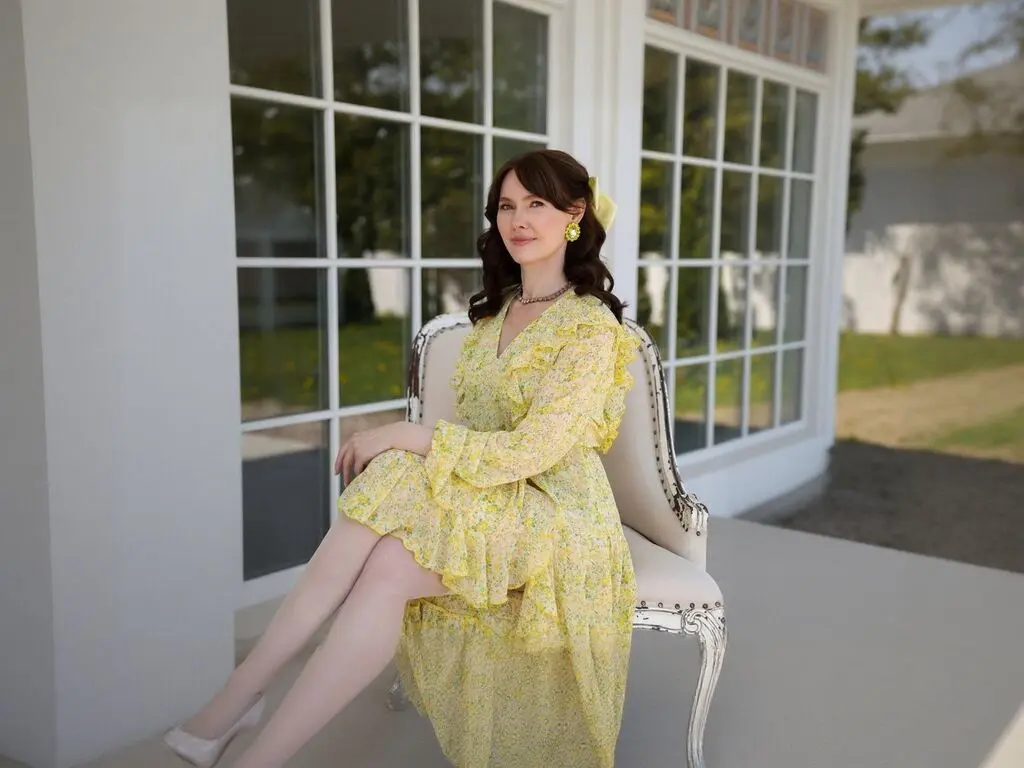 Elegant woman in a yellow floral midi dress sitting on a vintage chair by a bright window in a sunny setting.