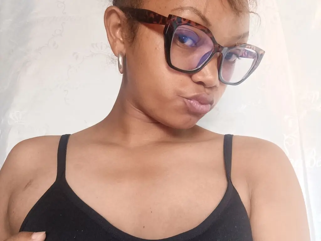 Young woman with glasses and black camisole posing in a bright indoor setting with a playful mood.